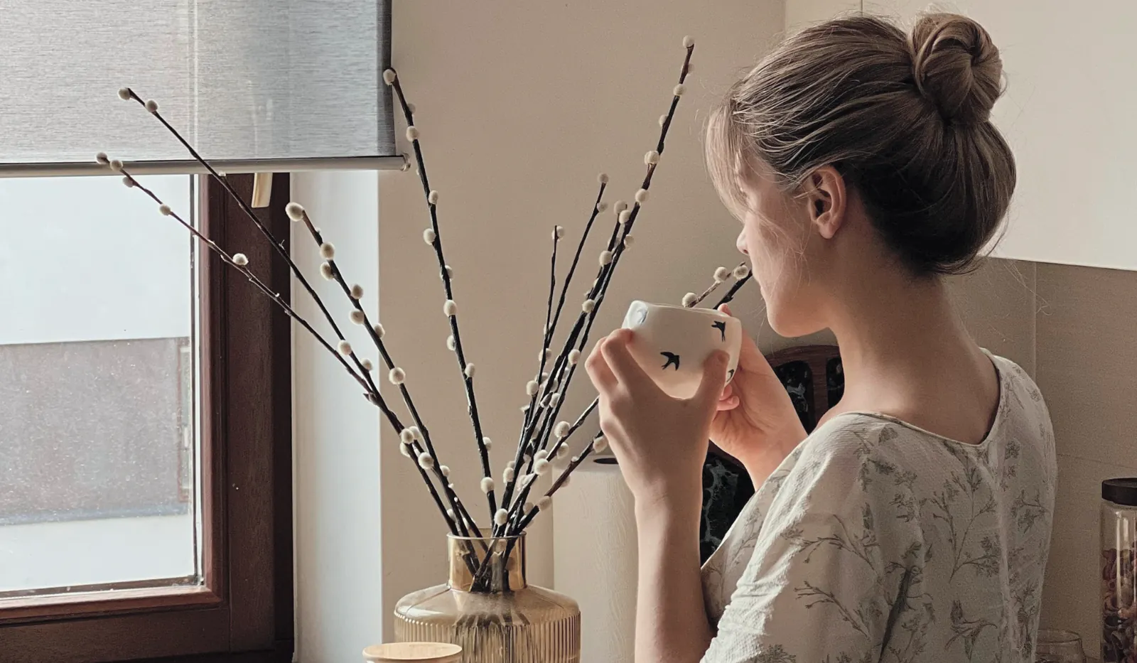 Een vrouw drinkt thee bij het keukenraam en kijkt naar buiten.